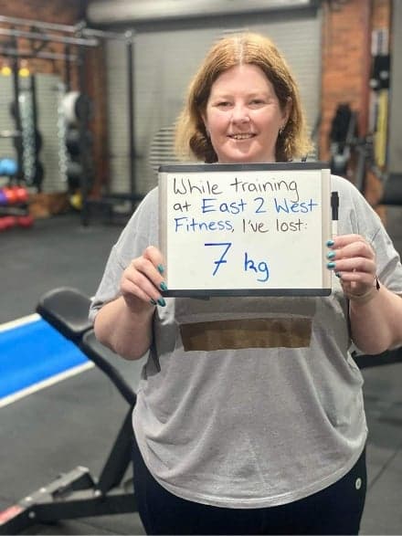 Client holding a whiteboard reading: while training at East 2 West Fitness I've lost 7 kg